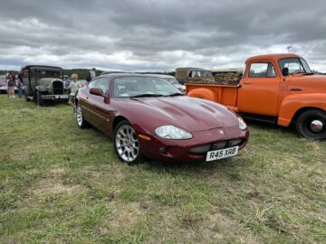 Een rode sportwagen met het kenteken "R45 XRB" staat geparkeerd op een grasveld, omringd door vintage voertuigen, waaronder een oude zwarte auto en een oranje vrachtwagen, onder een bewolkte hemel tijdens de West Oxon Steam & Vintage Show 2024.