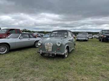 Een grijze vintage auto staat geparkeerd op een grasveld tussen andere klassieke voertuigen onder een bewolkte hemel tijdens de West Oxon Steam Vintage Show 2024.