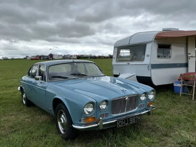 Een vintage blauwe auto met het kenteken "FDH 987K" staat geparkeerd op het gras naast een wit-blauwe caravan onder een bewolkte hemel tijdens de West Oxon Steam & Vintage Show 2024, perfect voor je FOTOVERSLAG.