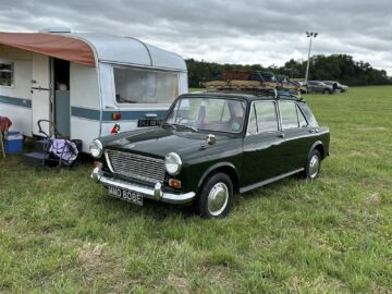 Een klassieke donkergroene auto geparkeerd op het gras naast een wit-blauwgroene caravan, vastgelegd tijdens de West Oxon Steam & Vintage Show 2024, met een bewolkte lucht op de achtergrond.