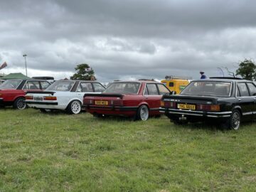 Verschillende oldtimers, waaronder modellen in rood, zwart en wit, staan geparkeerd in een grasveld onder een bewolkte hemel tijdens de West Oxon Steam & Vintage Show 2024. #FOTOVERSLAG