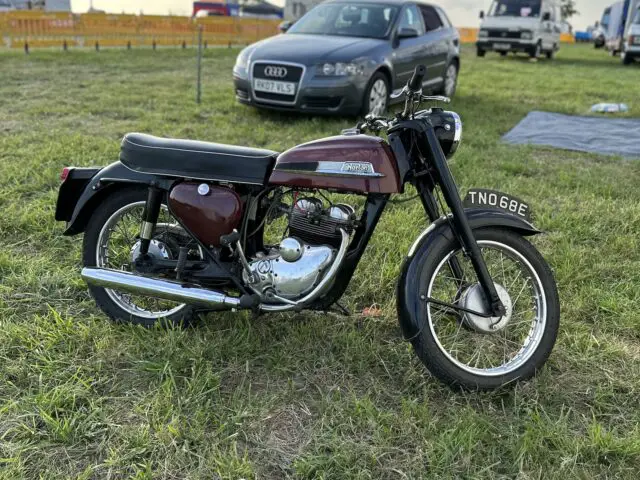 Vintage motorfiets geparkeerd op het gras, met een auto en tenten op de achtergrond tijdens de West Oxon Steam & Vintage Show 2024.