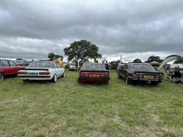 Op de West Oxon Steam & Vintage Show 2024 staan drie klassieke auto's geparkeerd op een grasveld onder een bewolkte hemel, van achteren gezien. Andere voertuigen en aanwezigen zijn zichtbaar op de achtergrond en vangen de tijdloze charme op een perfect FOTOVERSLAG-moment.