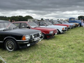 Een rij klassieke auto's, waaronder verschillende modellen in verschillende kleuren, staat geparkeerd op een grasveld onder een bewolkte hemel tijdens de West Oxon Steam & Vintage Show 2024.