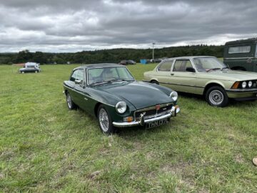 Een donkergroene klassieke auto met het kenteken "THN 137L" staat geparkeerd op een grasveld naast andere vintage voertuigen onder een bewolkte hemel tijdens de West Oxon Steam and Vintage Show 2024.