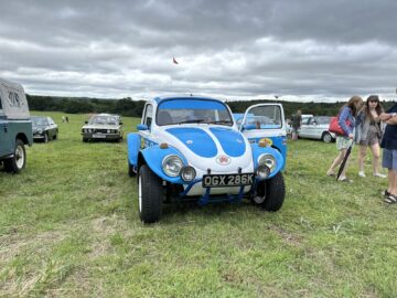 Een blauw-wit terreinwagen met kenteken "OGX 286K" staat geparkeerd op grasrijk terrein tijdens de West Oxon Steam & Vintage Show 2024. De deur aan de bestuurderszijde staat open en er staan verschillende mensen rond de auto en andere voertuigen op de achtergrond.