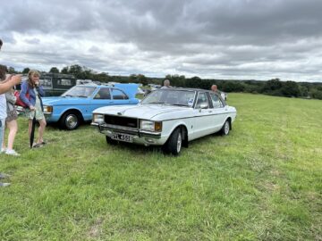 Een vintage witte auto met het kenteken "OYL 430R" staat geparkeerd op een grasveld, omringd door verschillende mensen en andere klassieke auto's onder een bewolkte hemel, en legt een moment vast uit het fotoverslag van de West Oxon Steam & Vintage Show uit 2024.