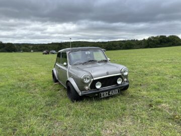 Een zilveren klassieke Mini-auto staat geparkeerd op een grasveld onder een bewolkte hemel tijdens de West Oxon Steam & Vintage Show 2024, met bomen en andere auto's op de verre achtergrond, perfect voor een fotoverslag.