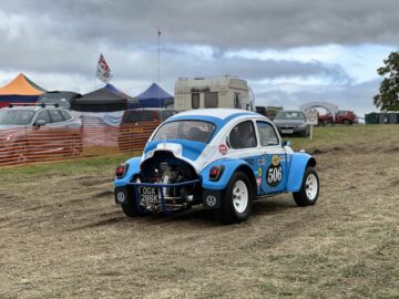 Een blauw-witte Volkswagen Kever met racenummer 506 staat geparkeerd op een grasveld nabij tenten en auto's, onder een bewolkte hemel op de West Oxon Steam & Vintage Show 2024. #FOTOVERSLAG