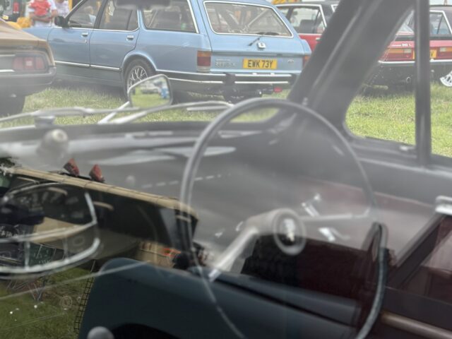 Binnen- en buitenaanzicht van klassieke auto's op de West Oxon Steam & Vintage Show 2024, gezien door het raam van een ander vintage voertuig. FOTOVERSLAG weet de nostalgische sfeer perfect vast te leggen.