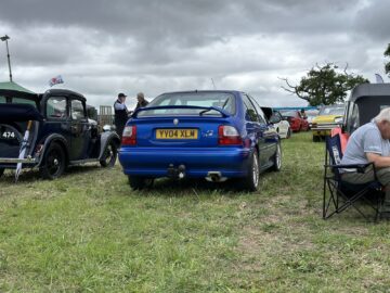 Een blauwe auto met kenteken "YY04 XLM" staat geparkeerd op gras, omringd door verschillende andere auto's en mensen op de achtergrond tijdens de West Oxon Steam & Vintage Show 2024. Rechts zit een oudere man op een klapstoel, waardoor een nostalgische sfeer ontstaat.