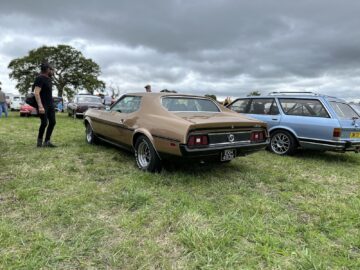 Een klassieke bruine auto staat geparkeerd op een grasveld bij de West Oxon Steam & Vintage Show 2024, met een paar mensen en andere voertuigen op de achtergrond onder een bewolkte hemel, wat de essentie van FOTOVERSLAG vastlegt.