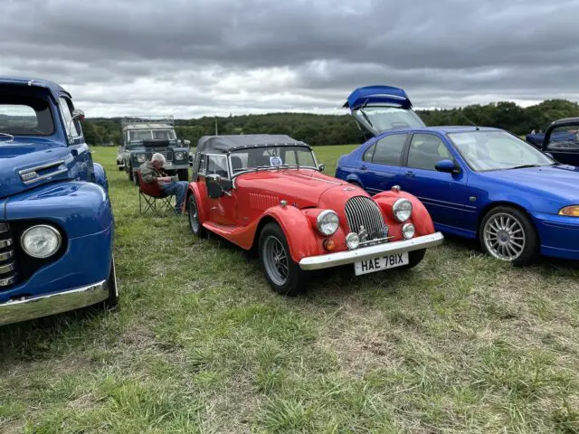 Een rij auto's op een grasveld tijdens de Vintage Show 2024, met een rode vintage cabriolet met kenteken HAE 781X, geflankeerd door een blauwe auto en een oudere blauwe vrachtwagen. Achter de vrachtwagen zit een persoon op een stoel.