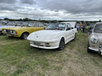 Op de West Oxon Steam & Vintage Show 2024 staat een klassieke witte auto met kenteken JBW 601Y geparkeerd op gras, omringd door andere oldtimers en bezoekers.