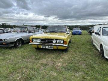 Een gele klassieke auto met kenteken "JVB 184N" staat geparkeerd op gras, omringd door andere klassieke auto's, onder een bewolkte hemel tijdens de West Oxon Steam & Vintage Show 2024. #FOTOVERSLAG