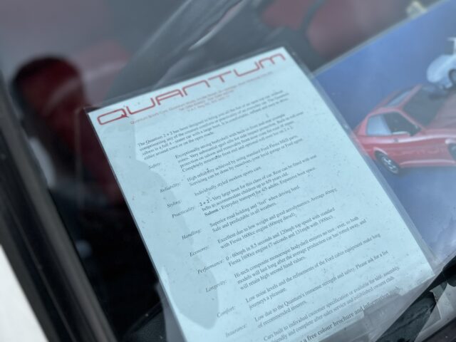 Een document met de titel "Quantum", verwijzend naar de West Oxon Steam & Vintage Show van 2024, wordt op het dashboard van een auto geplaatst, zichtbaar door de voorruit. Het document bevat een lijst met kenmerken en specificaties. Ernaast is gedeeltelijk een kleurrijke brochure zichtbaar.