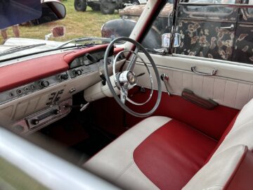 De afbeelding toont het interieur van een klassieke auto op de West Oxon Steam & Vintage Show 2024, met rood-wit gestoffeerde stoelen, een wit stuur, een rood dashboard en verschillende bedieningsknoppen en draaiknoppen.