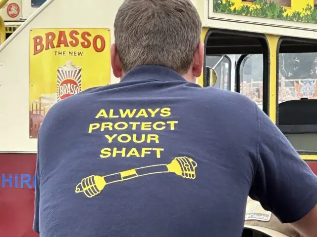 Een man met kort haar zit met zijn rug naar de camera en draagt een blauw shirt met de gele tekst "ALWAYS PROTECT YOUR SHAFT" voor een posterdisplay in vintage-stijl ter promotie van de West Oxon Steam Vintage Show 2024.