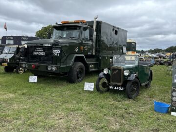 Een groot militair voertuig met het label "Long Wheelbase Unipower" staat geparkeerd naast een oldtimer met kenteken "WV 4432" op een grasveld onder een bewolkte hemel tijdens de West Oxon Steam Vintage Show 2024.