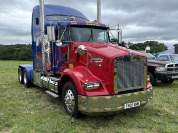 Een rood-blauwe Kenworth semi-truck met chromen details en dubbele uitlaatpijpen staat geparkeerd op grasrijk terrein onder een bewolkte hemel tijdens de West Oxon Steam & Vintage Show 2024.