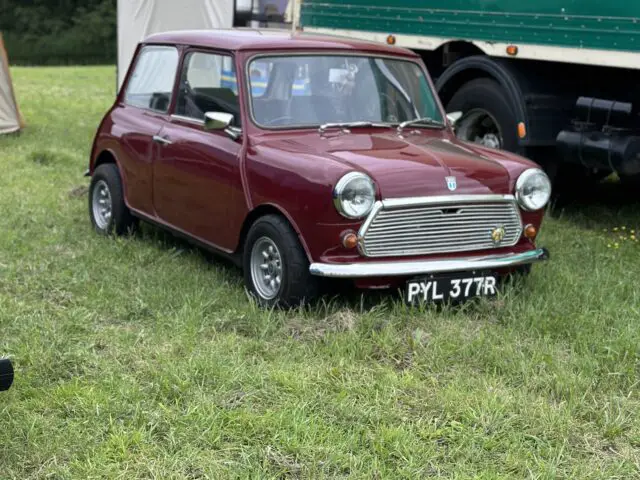 Een klassieke rode mini-auto met een wit dak staat geparkeerd op gras naast een groot wit en groen voertuig tijdens de West Oxon Steam & Vintage Show 2024, met een kleine tent op de achtergrond, perfect voor een FOTOVERSLAG-functie.