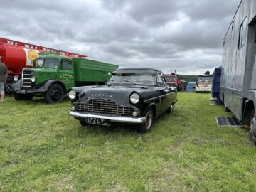 Een klassieke zwarte auto, kenteken 172 UYL, wordt tentoongesteld op een grasveld tijdens de West Oxon Steam & Vintage Show 2024. Op de achtergrond zijn andere vintage vrachtwagens en auto's zichtbaar, die de essentie van het FOTOVERSLAG-evenement van dit jaar vastleggen.