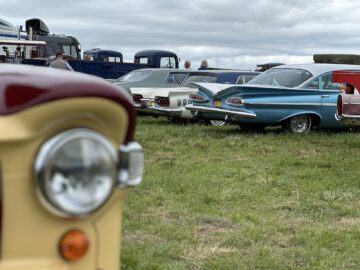 Oldtimers geparkeerd op een grasveld, met de koplamp van een klassieke beige auto op de voorgrond en verschillende andere klassieke auto's, waaronder blauw-witte modellen, zichtbaar op de achtergrond tijdens de West Oxon Steam Vintage Show 2024.