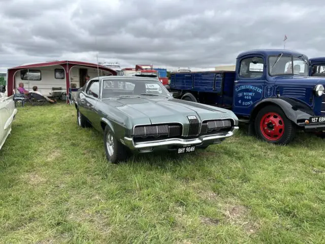 Een vintage groene cabriolet staat geparkeerd op het gras naast een caravan en een blauwe klassieke vrachtwagen onder een bewolkte hemel, wat perfect de essentie van de West Oxon Steam & Vintage Show 2024 weergeeft voor een komende FOTOVERSLAG.