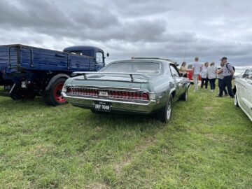 Een lichtgroene klassieke auto met licentie BNY 904H staat geparkeerd op het gras naast een blauwe vrachtwagen tijdens de West Oxon Steam & Vintage Show 2024. Er staan verschillende mensen omheen, met een bewolkte hemel boven hun hoofd, die momenten vastleggen voor een fotoverslag.