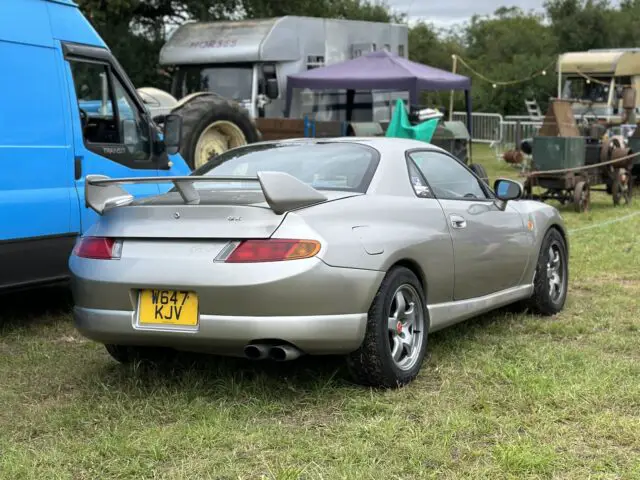 Een zilveren sportwagen met een achterspoiler en geel kenteken, geparkeerd op gras nabij een blauw busje en diverse uitrusting op de West Oxon Steam & Vintage Show 2024.