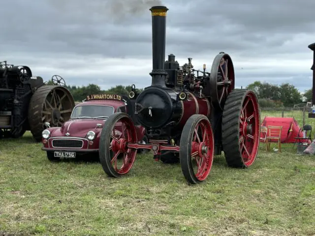 Een vintage stoomtractor met rode wielen en een verticale schoorsteen staat geparkeerd naast een kleine klassieke auto, beide op een grasveld onder een bewolkte hemel tijdens de West Oxon Steam & Vintage Show 2024.