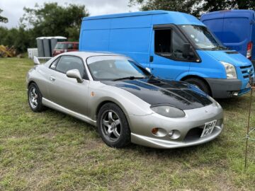 Op het gras staat een zilveren sportcoupé met zwarte motorkap geparkeerd, naast een blauw busje. Bomen en andere voertuigen bevinden zich op de achtergrond en vangen een stukje van de West Oxon Steam & Vintage Show 2024 op.