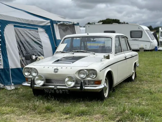 Een klassieke witte auto staat geparkeerd op het gras naast een blauw-witte tent op de West Oxon Steam & Vintage Show 2024, met caravans op de achtergrond onder een bewolkte hemel. (FOTOVERSLAG)